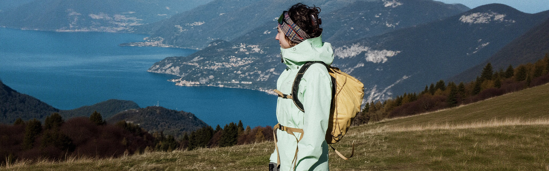 Person with a backpack standing on a grassy hill overlooking a lake and mountains.